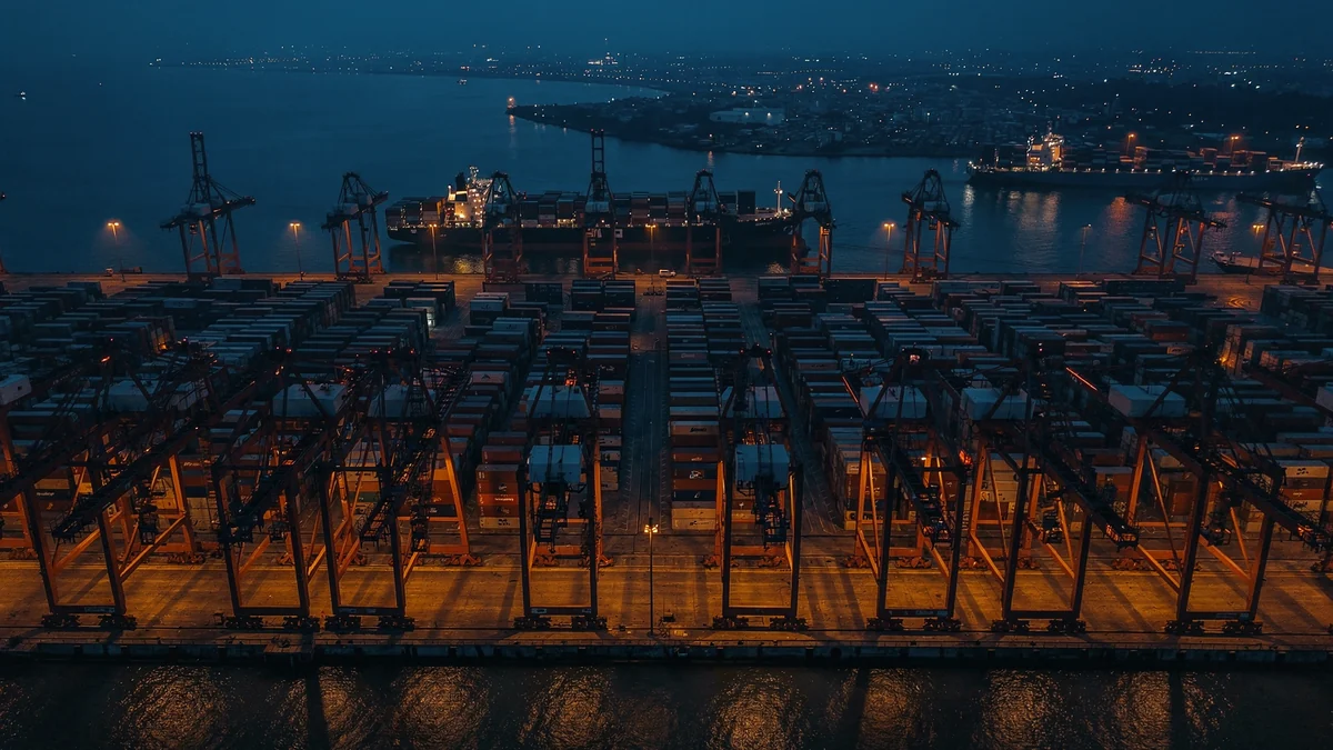 Aerial view of a container terminal at Port Klang at dusk, with rows of ship-to-shore gantry cranes and stacked containers alongside a berthed container vessel